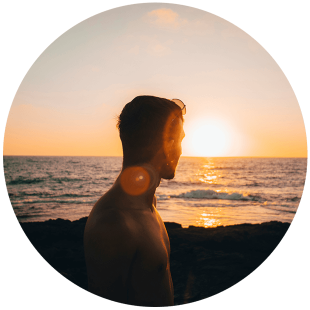 Silhouetted man standing shirtless on a rocky beach, gazing toward the ocean as the sun sets on the horizon, casting a warm golden glow.